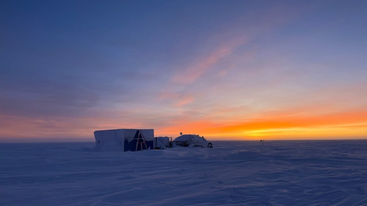 A lone square building with small structures sitting a vast, flat area of snow during twilight. 