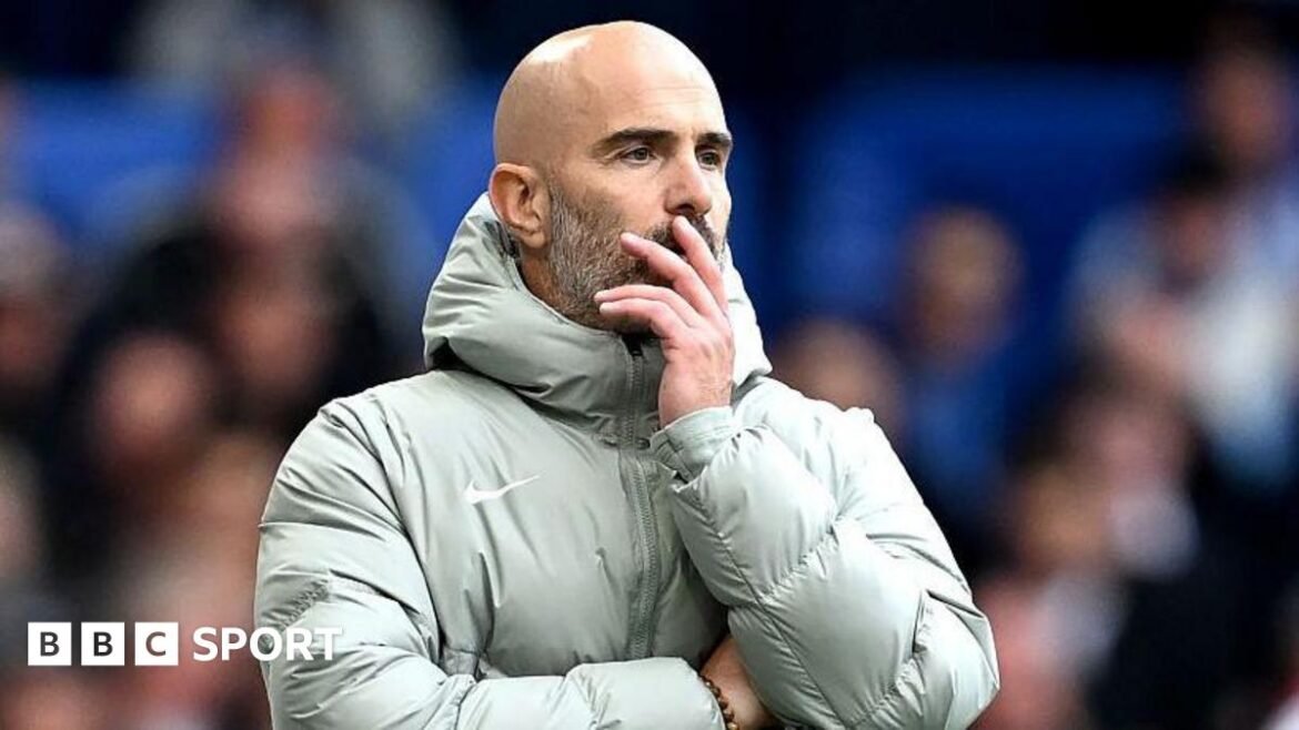 Enzo Maresca, looks on during the Premier League match between Chelsea and Brighton at Stamford Bridge. He is wearing a grey coat and holding his face.
