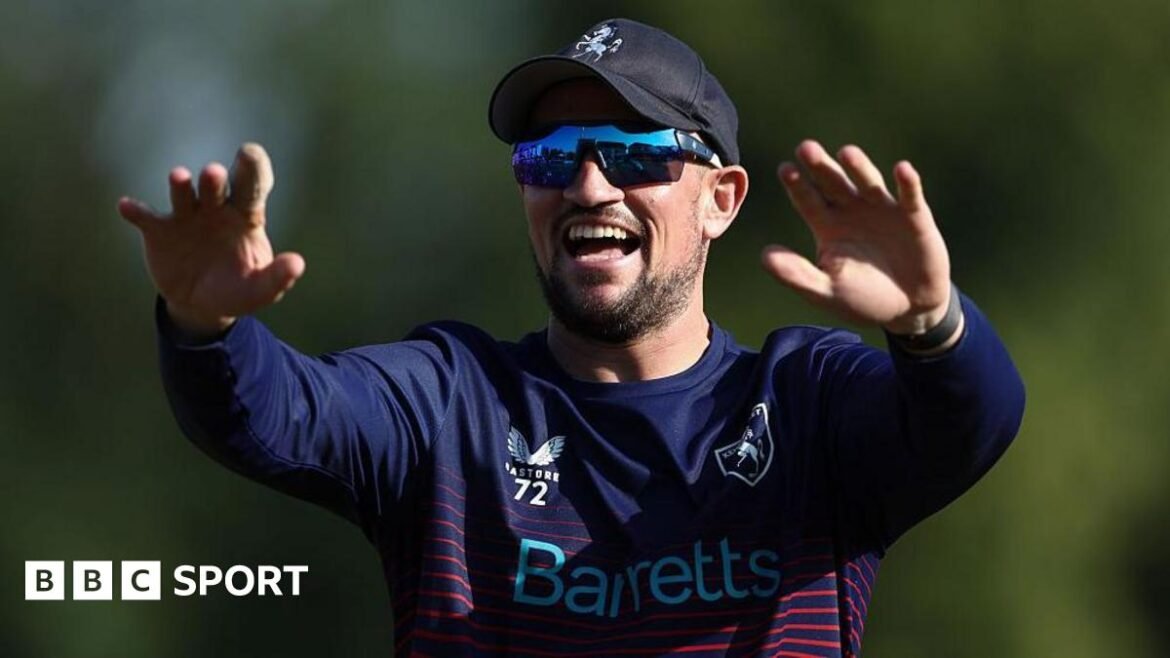 Harry Finch smiles with both arms raised in front of him, wearing a blue baseball cap, dark sunglasses and a blue cricket kit