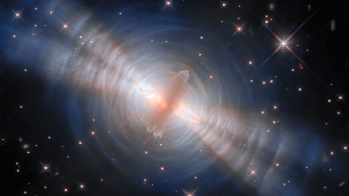 An image of the egg nebula, with a glowing streak of pink gas in the middle surrounded by concentric circles of white like with four diagonal beams of light streaking from top left to bottom right all in front of a deep space starry background