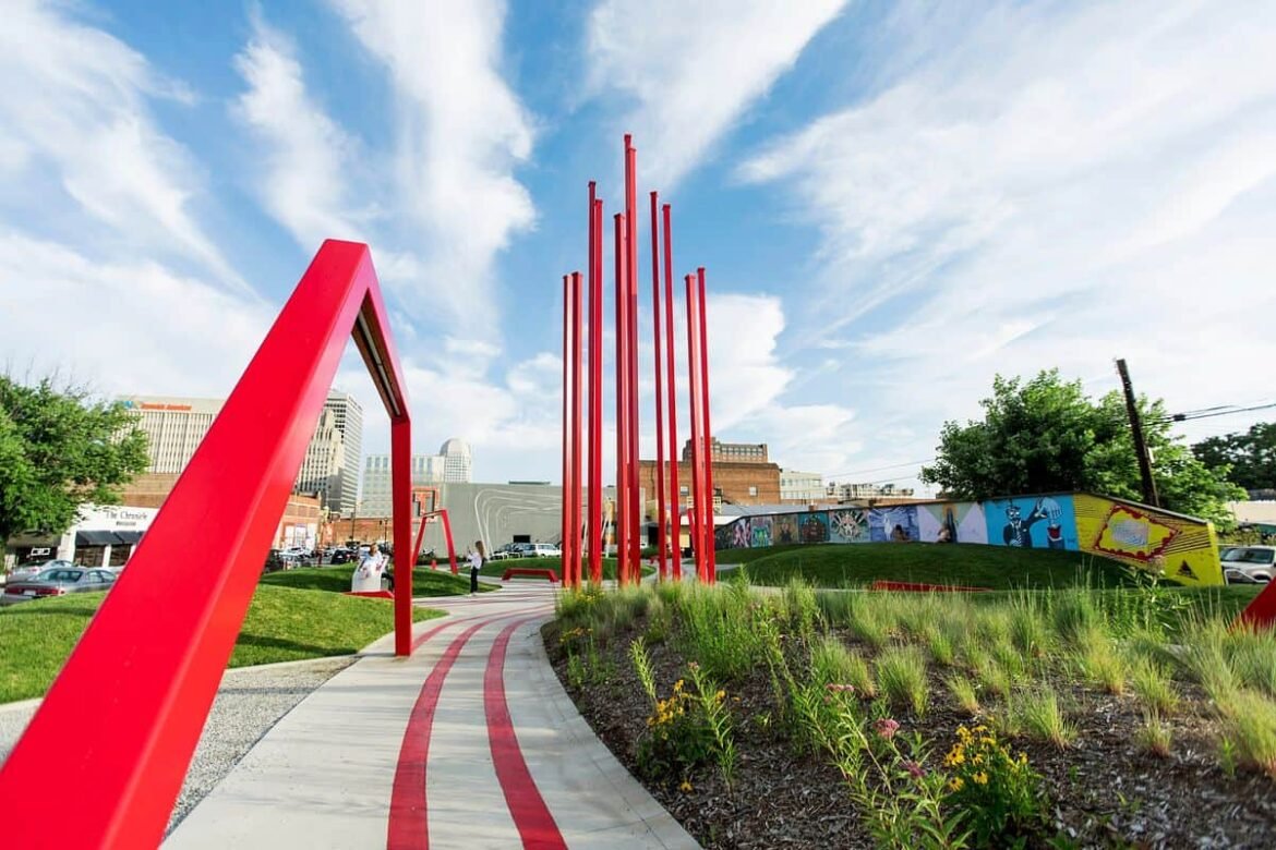 Landscape shot of ARTivity on the Green in Winston-Salem during daytime. Blue skies and greenery with flowers in bloom. Red sculptures in foreground with city in background. Spring or summer season.