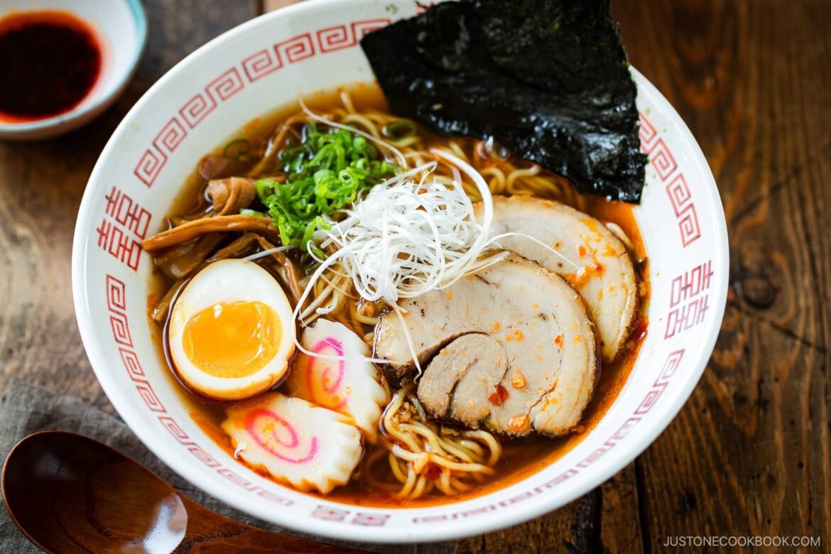 A bowl of spicy shoyu ramen topped with sliced pork, half a soft-boiled egg, green onions, seaweed, bamboo shoots, fish cake slices, and white shredded onions, served in a patterned bowl on a wooden table.
