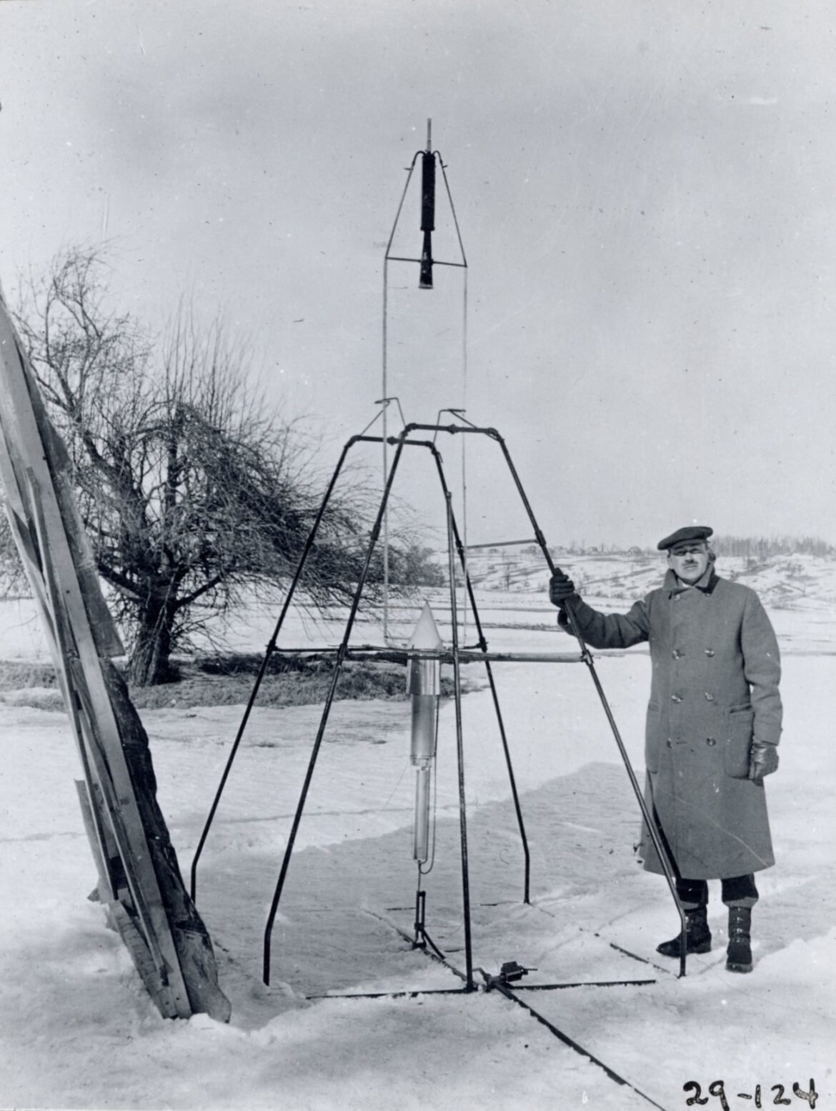 On a snowy March 16, 1926, Dr. Robert H. Goddard rests his hand on the testing frame supporting his liquid fuel rocket at Ward Farm in Auburn, Massachusetts. A wooden door is propped up at an angle next to the frame where Goddard’s assistant, Henry Sachs, later sheltered after lighting the rocket.