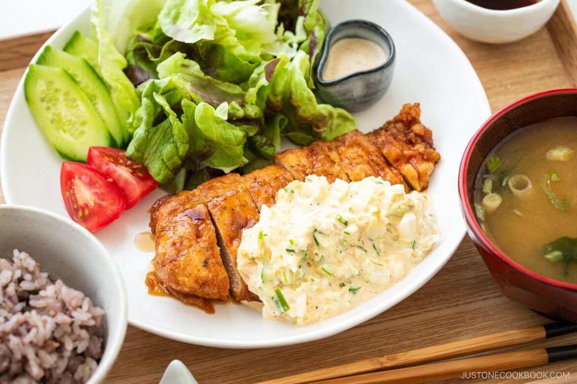 A plate of crispy chicken nanban topped with creamy egg salad, served alongside fresh lettuce, cucumber, tomato, a small dish of sauce, a bowl of miso soup, and multigrain rice.