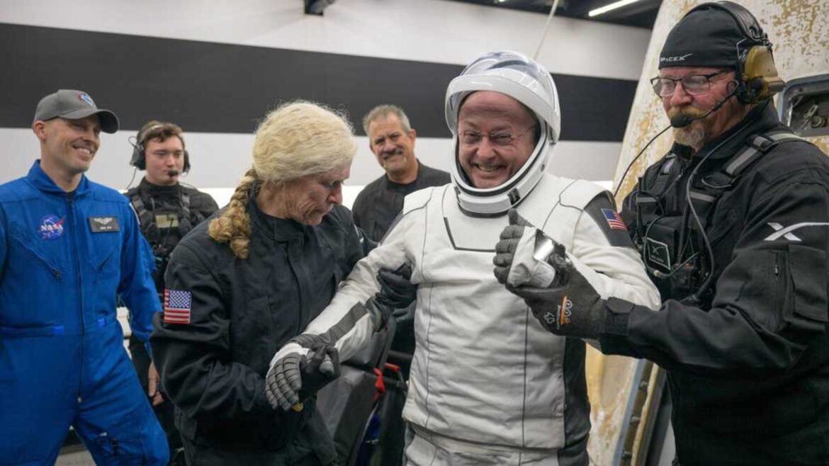 A man wearing a white spacesuit is accompanied by two people wearing black jumpsuits.