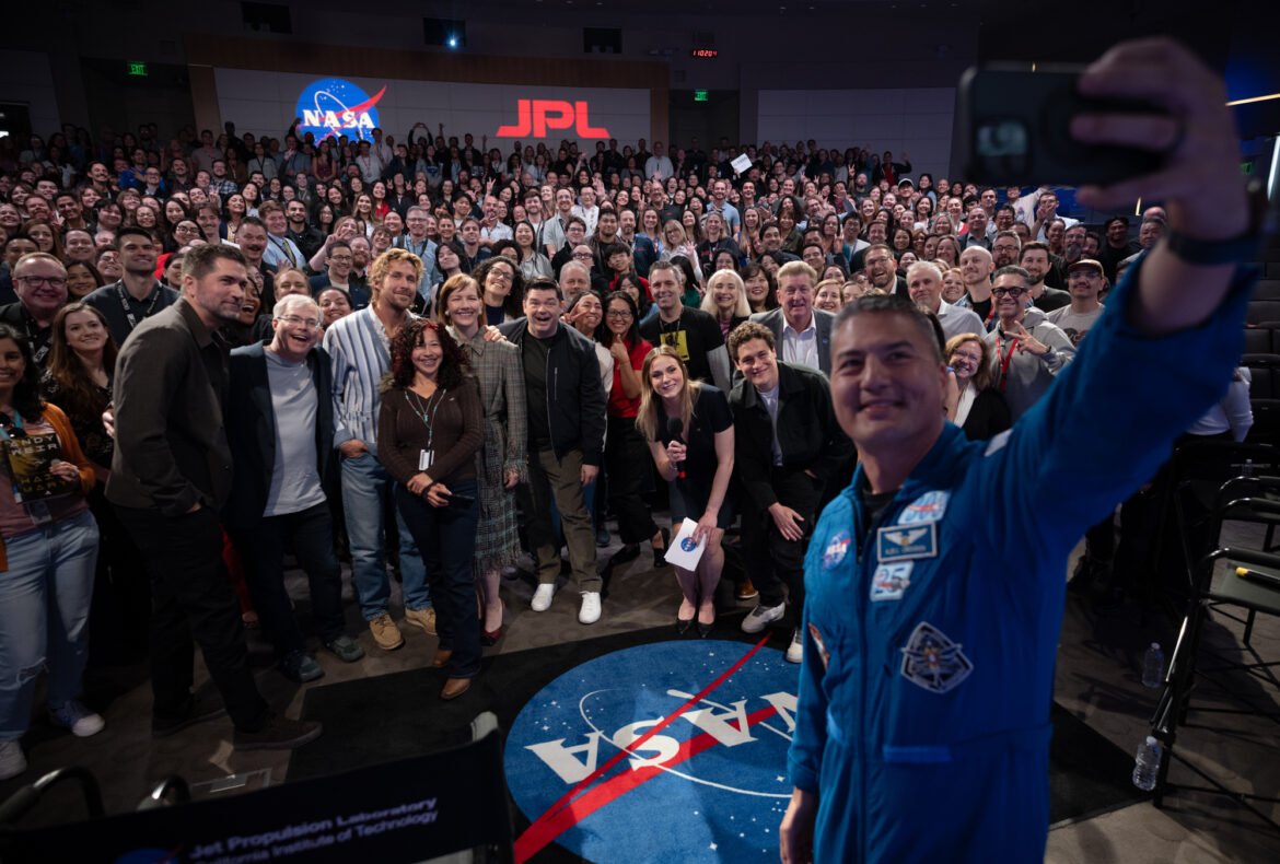 At right, a man in a blue flight suit with patches on it takes a selfie with his cell phone. He is in the photo, as well as panel members and the large audience. They are in an auditorium. In the far background, there is a NASA "meatball" insignia and the letters "JPL" on the wall.