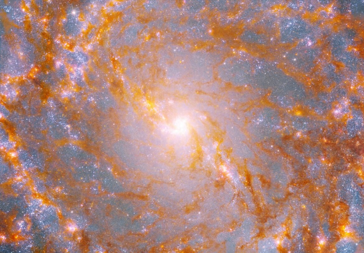 A close-up view shows a galaxy’s bright, yellow center, surrounded by patches of glowing orange-brown dust and pink stars.