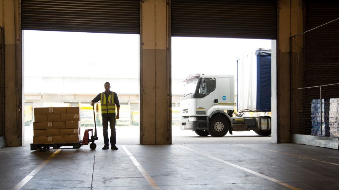 A worker wearing a yellow vest stands looking into an empty warehouse with a cart full of food boxes next to him and a truck in the background