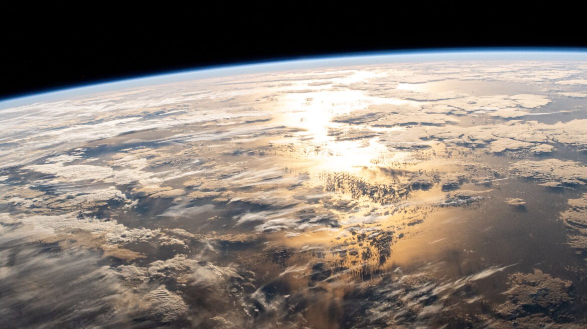 A view of the Atlantic Ocean from the International Space Station. Sun shines on the water, turning the water in the middle of the photo yellow-orange. There are flat, white clouds scattered above the ocean. In the distance, Earth’s gentle curve is outlined by a pale blue hazy line – that’s our atmosphere. Beyond that is the darkness of space.