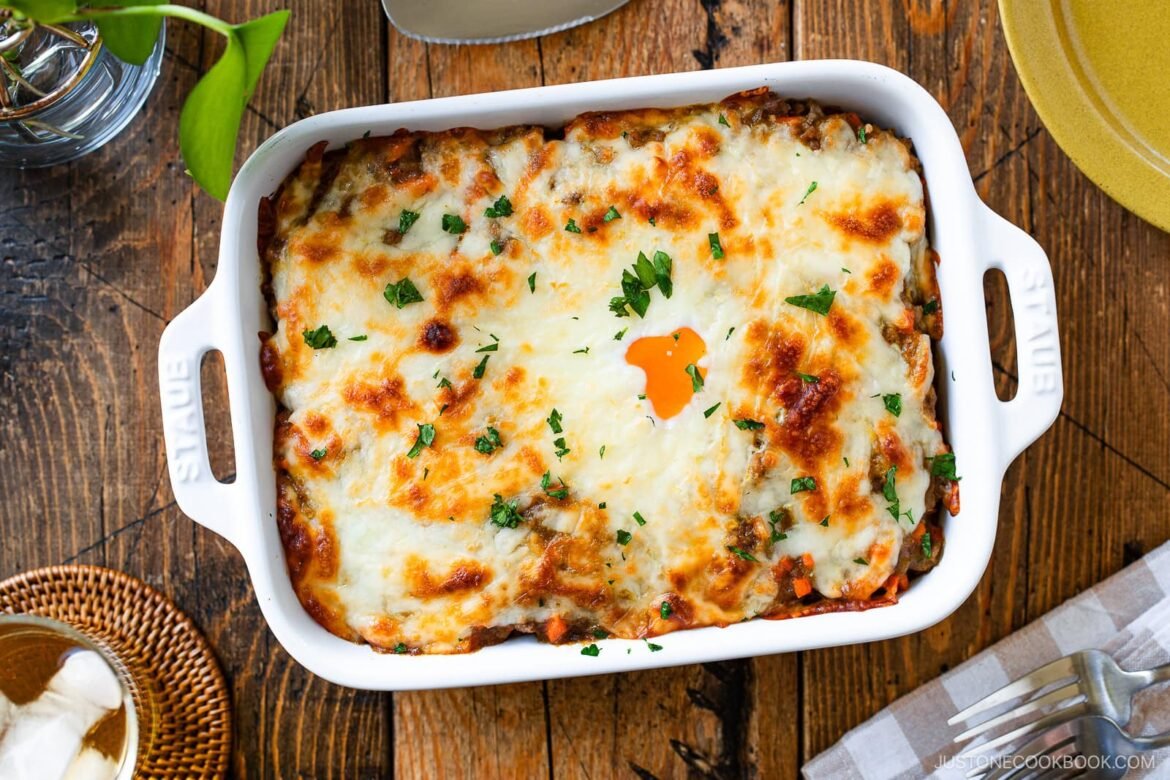 A baked Yaki Keema Curry casserole topped with melted cheese and parsley sits in a white dish on a wooden table, surrounded by plates, utensils, a napkin, and a glass of water.