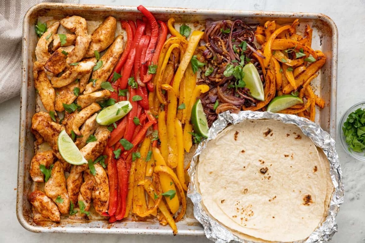 Baked sheet pan chicken fajitas with bell peppers, garnished with cilantro and lime wedges with a foil pack of tortillas on tray.
