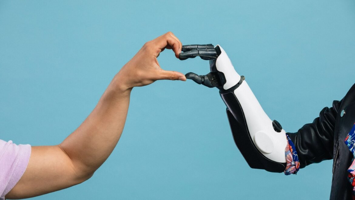 A person's left hand comes from left of the image to meet a black and white robotic hand from the right of the image to make a heart with their hands in the center, all in front of a blue background.