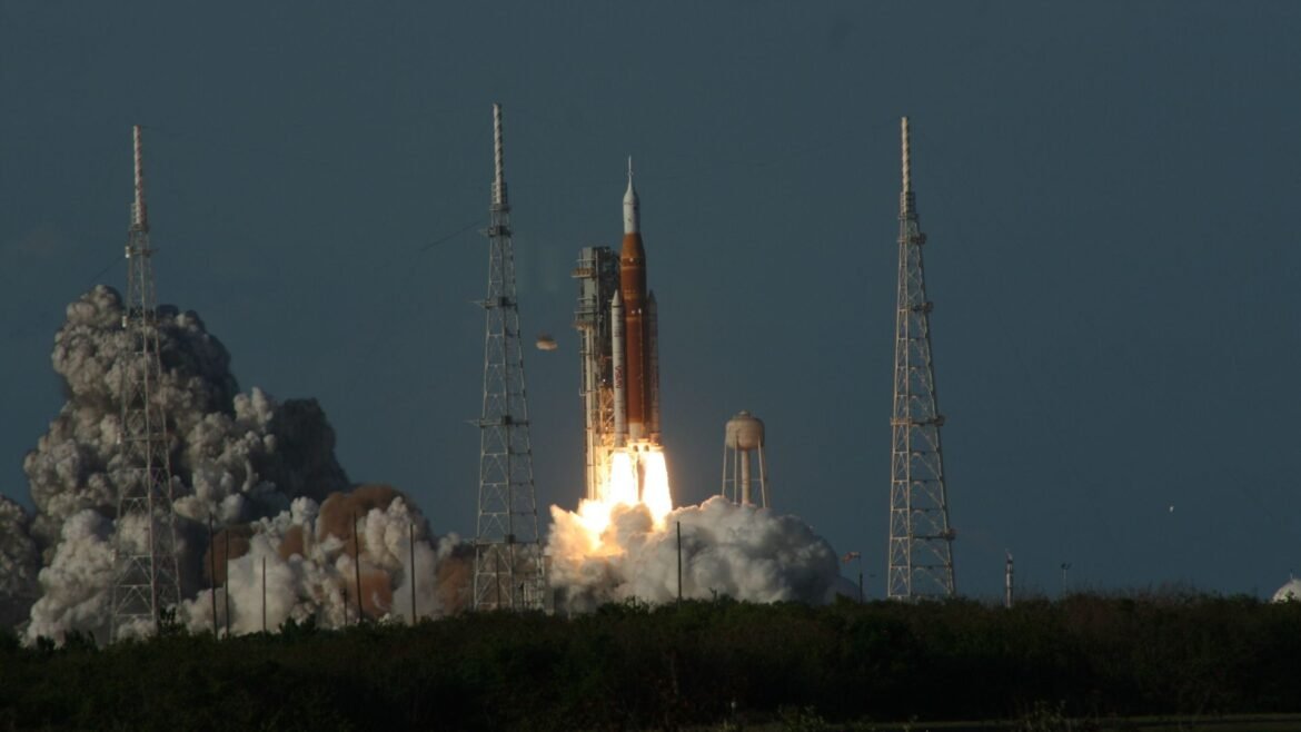 An orange and white rocket blasts off a launchpad, billowing gray smoke behind it