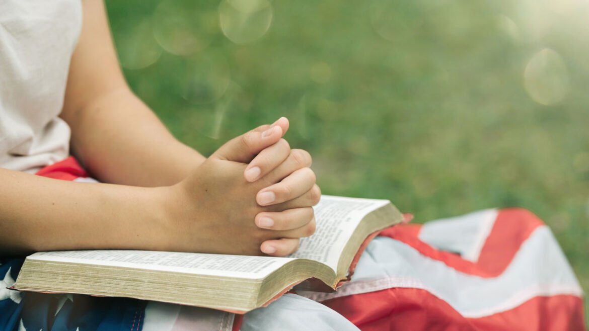 Close-up of young person with flag and Bible on their lap in outdoor setting.