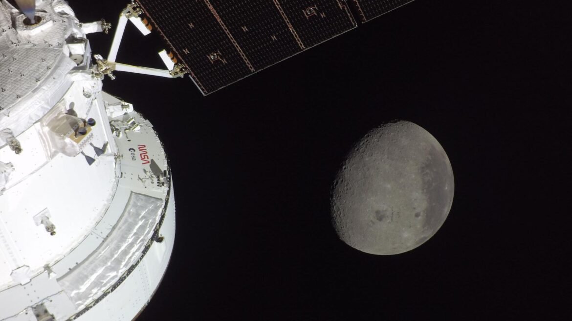 A gray sphere in the darkness of space is seen from a white spacecraft with solar panels to the left of the image. 