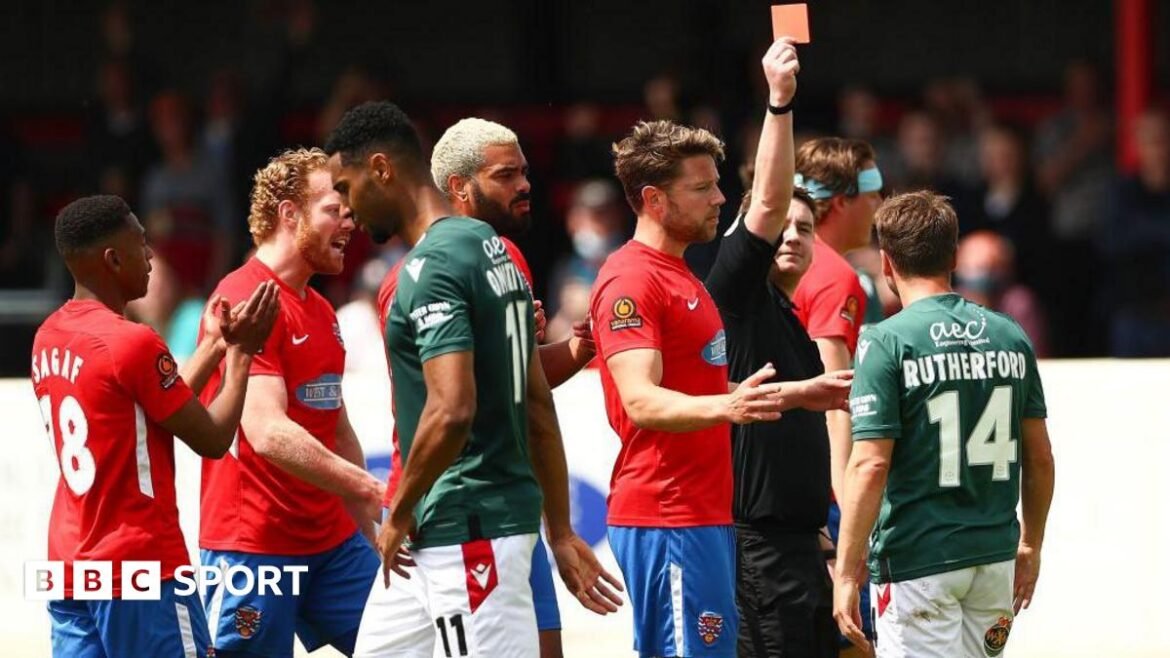 Paul Rutherford is shown a red card with his back to the camera showing his name and number in Wrexham's green away shirt, with Dagenham players in red shirts around the referee.