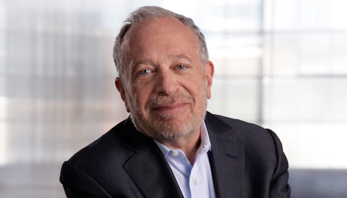 informal horizontal portrait of Robert Reich, wearing a blazer and a light blue shirt, against a bright silver-white background