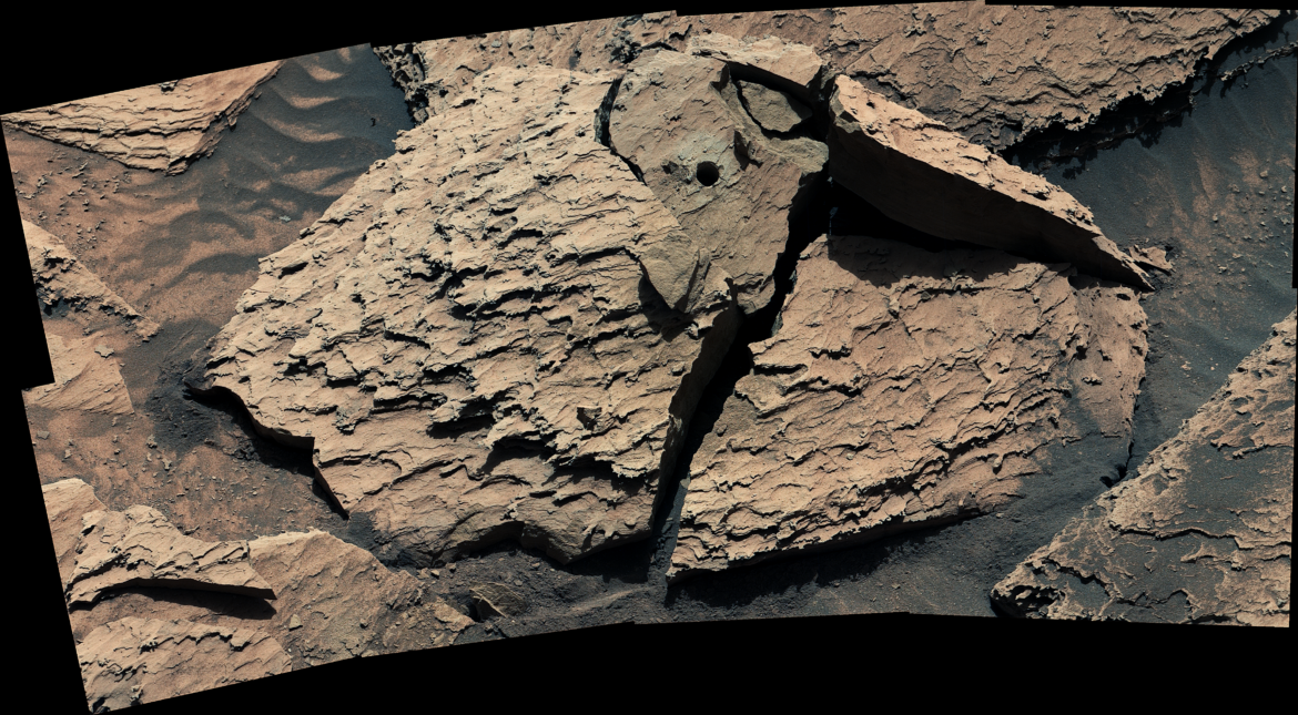 A large, reddish-brown rock as seen by NASA's Curiosity Mars rover. The rock is broken into several pieces. A drill hole is visible in the top middle piece.