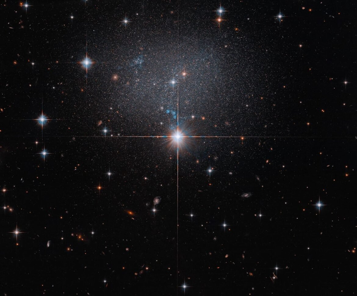 A star in the foreground shines brightly with four diffraction spikes. Above this star is a diffuse and faint dwarf irregular galaxy. The galaxy itself has bright blue and red stars.