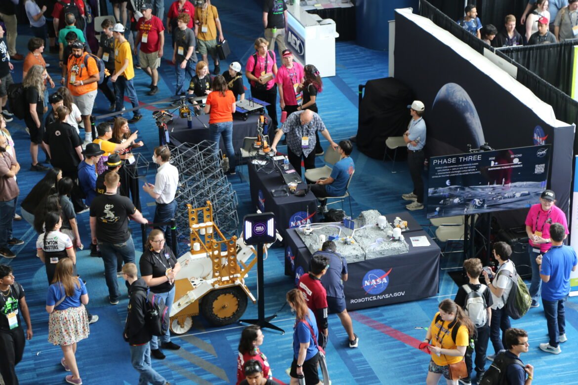 Birds-eye view of a NASA exhibit at the FIRST Robotics Championship in Houston, with a model of NASA's Moon Base and a lunar sampling cart in the foreground.