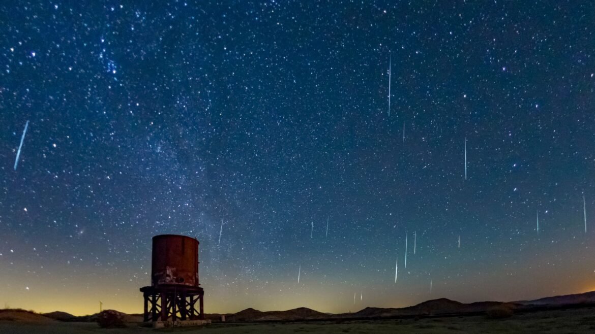 A dark blue night sky is streaked with white meteors moving through space with a rusty cylindrical water tower below.
