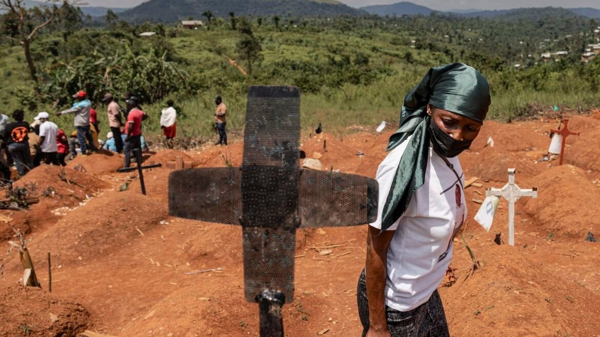  A woman wearing a mask walks by a grave marker with other people in the background