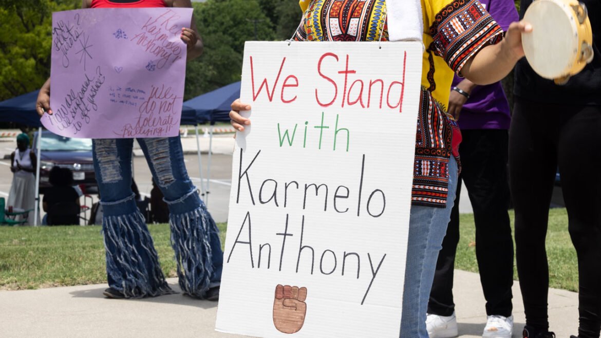 Protester in African garb holds sign reading "We Stand With Karmelo Anthony."