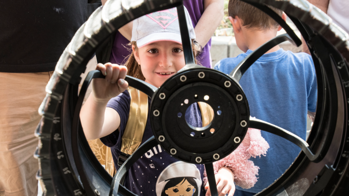 A smiling young girl looking through the spokes of a large black metal wheel.