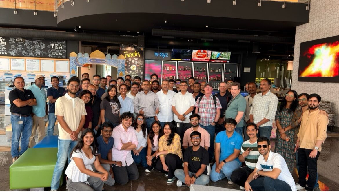 A large group of employees and interns pose together inside a modern office café/lounge area. The group includes about 40 people standing and kneeling in several rows. Behind them are coffee and tea menu boards, colorful digital displays, and a decorative Ramadan-themed arch. The space features bright lighting, contemporary furniture, and an open, collaborative atmosphere.