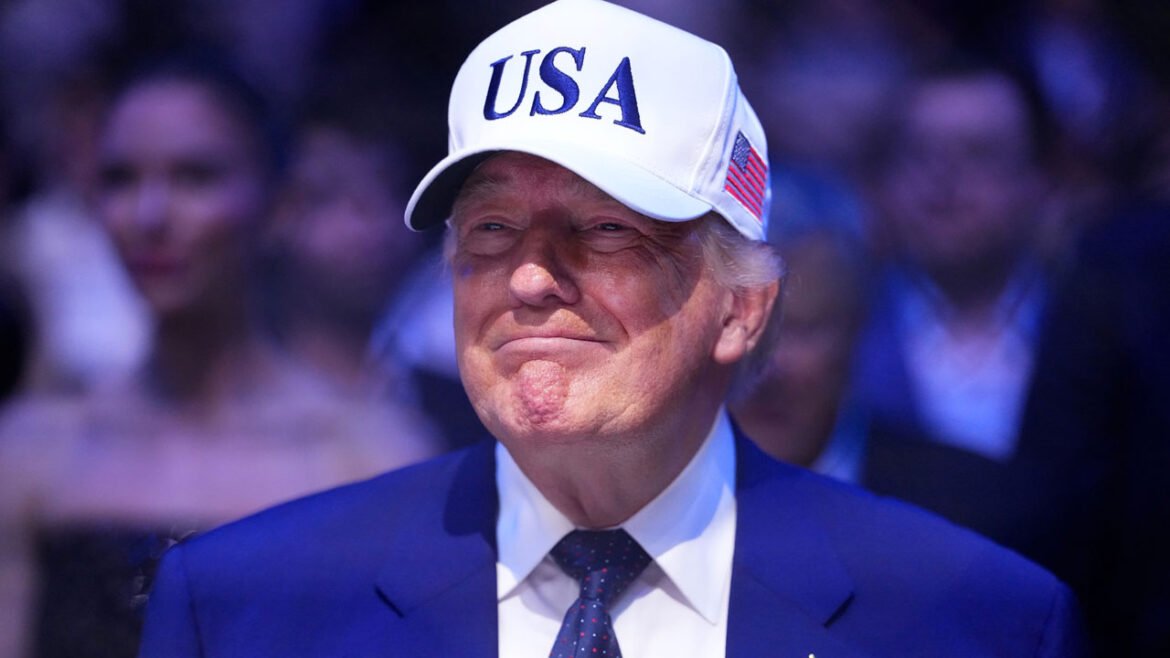 A grinning Donald Trump in a blue suit with white baseball cap with USA in blue lettering.