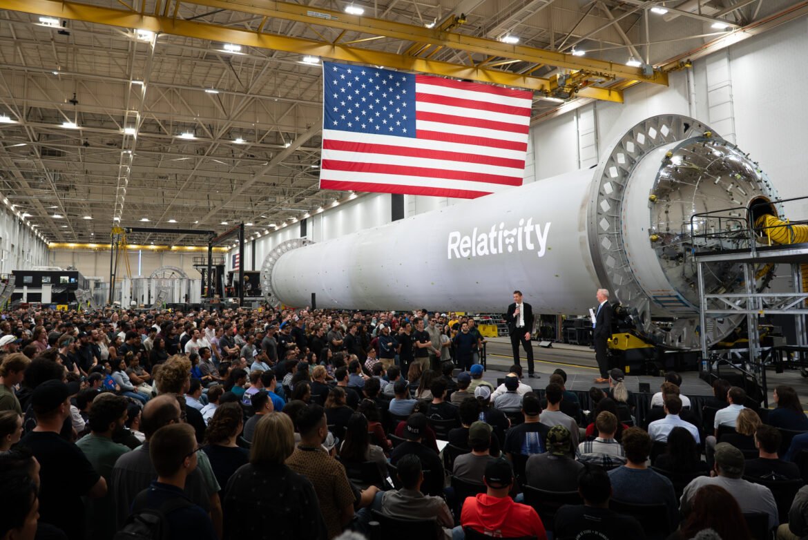 NASA Administrator Jared Isaacman stands in front of a large, cylindrical, gray spacecraft with a white Relativity logo on it and addresses hundreds of Relativity Space employees. A large American flag hangs from the ceiling.