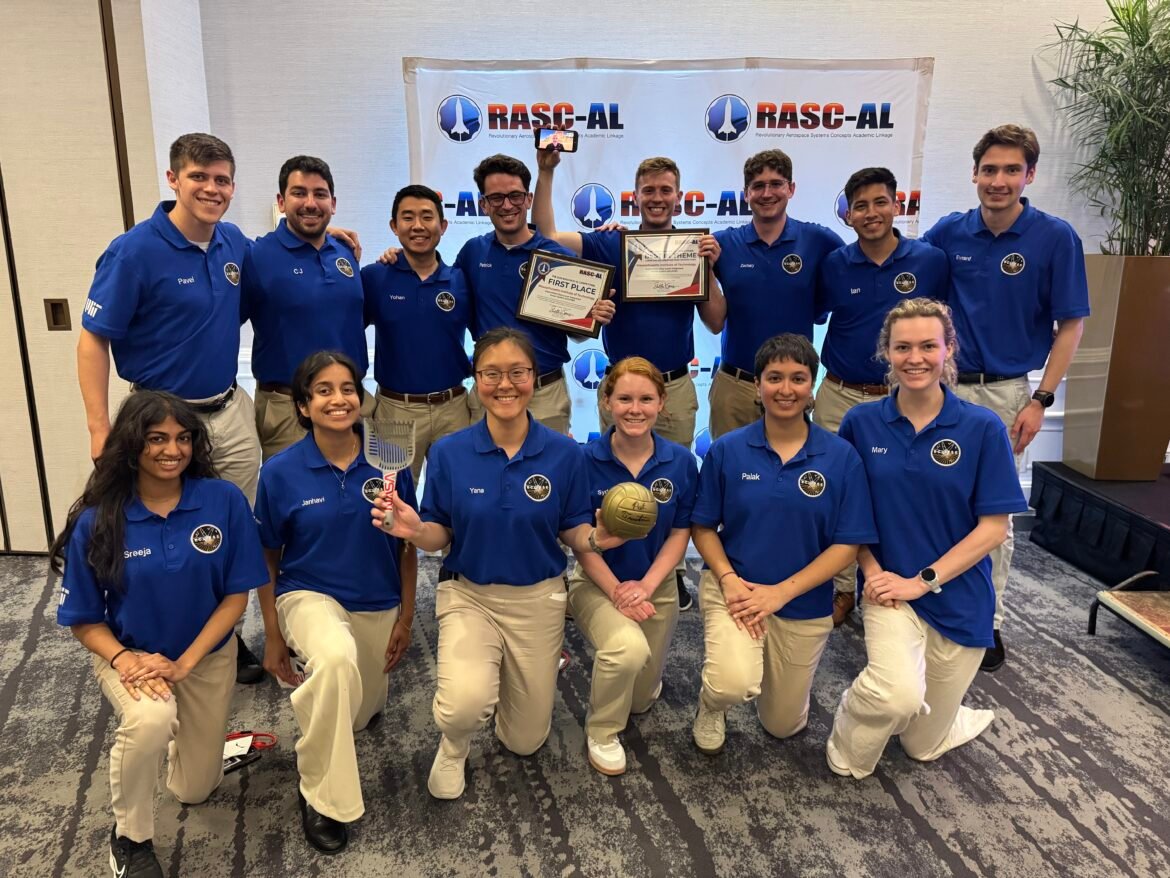 A group of 14 students wearing blue polo shirts smile at the camera while holding the first place certificate for winning the NASA RASC-AL challenge.