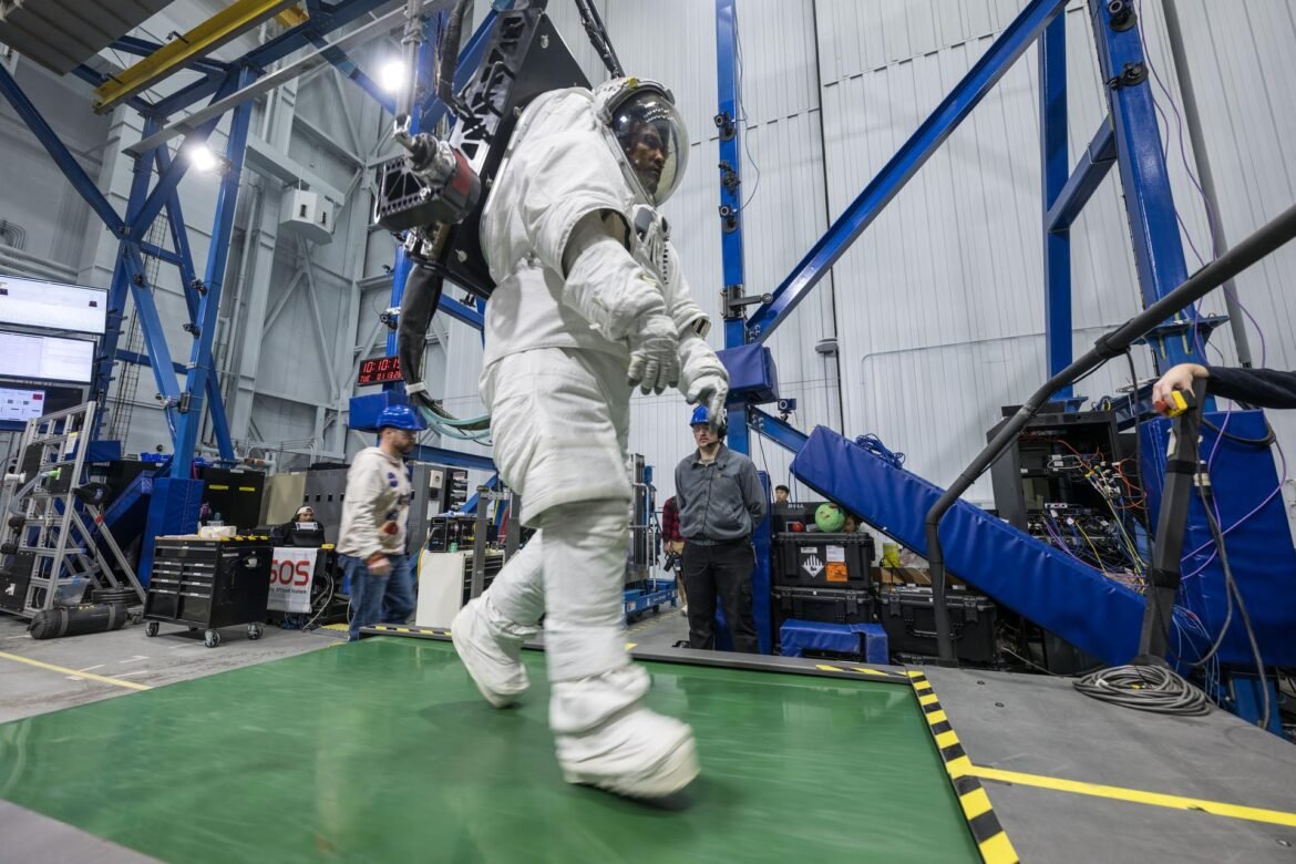 Dressed in a white space suit—with hands gloved, feet booted and head in a helmet with a clear visor—Artemis II astronaut Victor Glover walks on a wide green treadmill. His suit is harnessed to a black machine that is taking the weight off Glover such that, while in the suit, he experiences forces equivalent to lunar gravity. Two people wearing casual clothes and blue hard hats stand in the background, waiting to support Glover and the experiment, should the need arise.  Behind Glover are crisscrossing blue iron beams that stretch to a high ceiling in this industrial building at NASA’s Johnson Space Center.