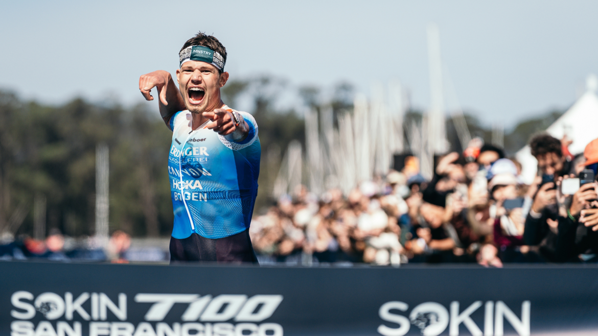 German pro triathlete celebrates his T100 victory as he runs down the finish chute toward the tape with spectators cheering him on outside on sunny day