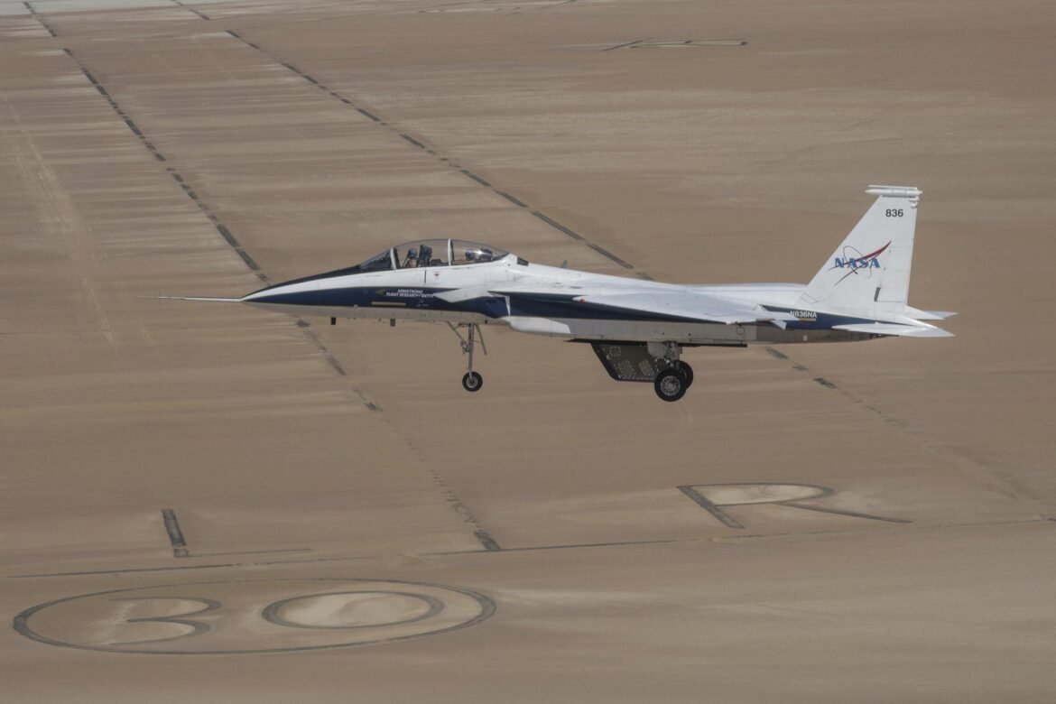 A white and blue painted jet aircraft flies above a tan lakebed with the gear and wheels out. A model wing hangs below the aircraft’s center line. The cockpit is closed and two pilots are visible inside with flight gear on.