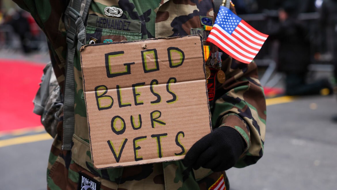 Individual in military fatigues holds a small flag and a cardboard sign reading "God Bless Our Vets."