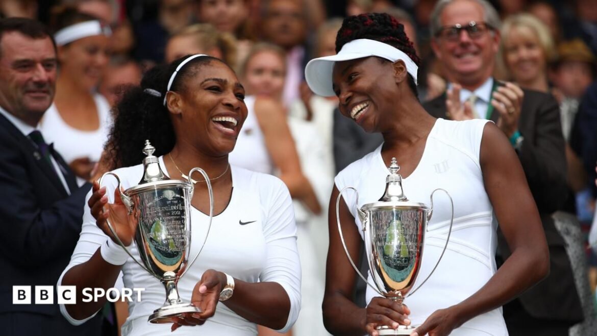 Venus Williams, in short white top and cap, stands alongside Serena Williams, who wears a black outfit