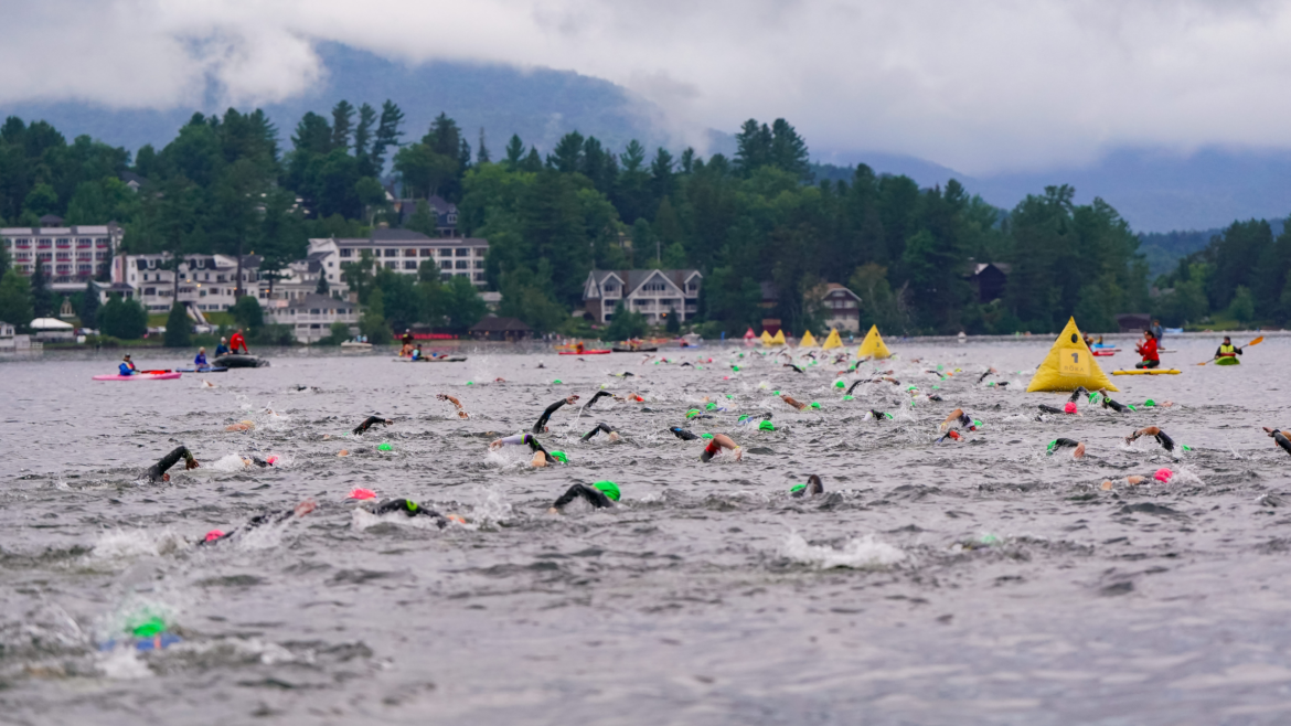 dozens of triathletes in wetsuits and swim caps swimming in Mirror Lake at Ironman Lake Placid