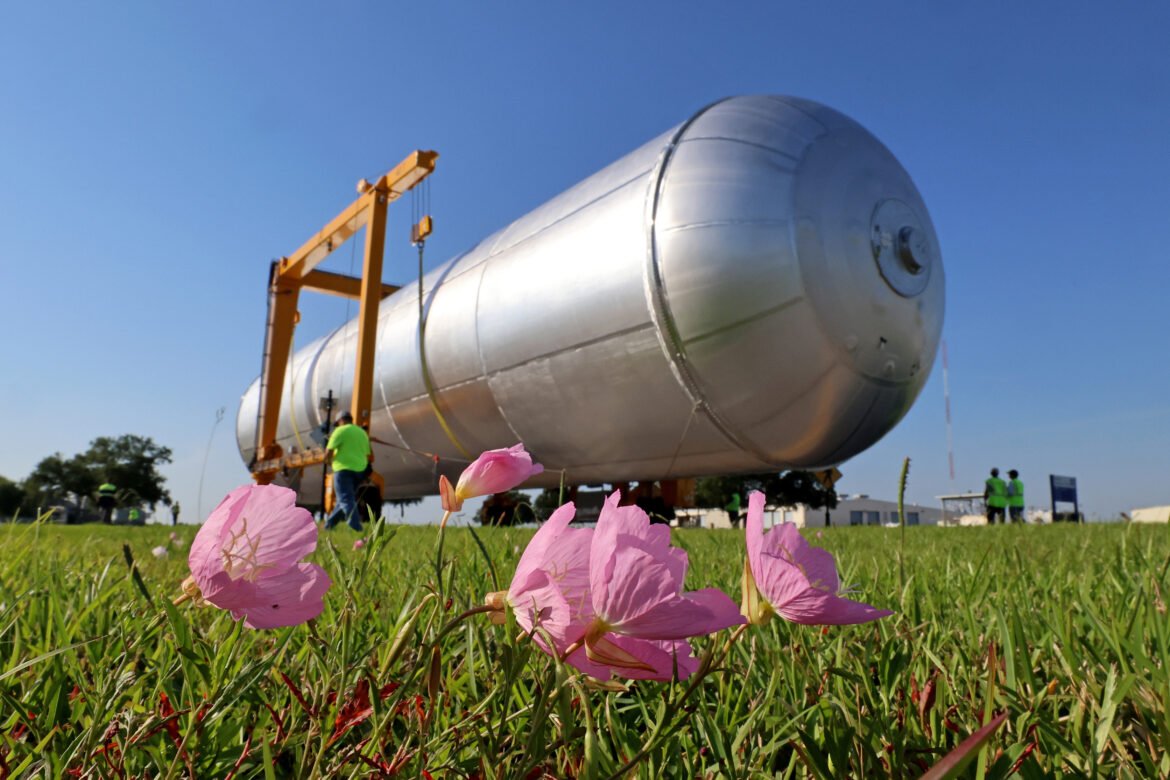 In the foreground, a few pink flowers on a grassy field lean to the right. Behind them is a long, silver cylinder being moved by crews in bright green safety shirts and vests. The massive object is the liquid hydrogen tank for the Artemis IV rocket.