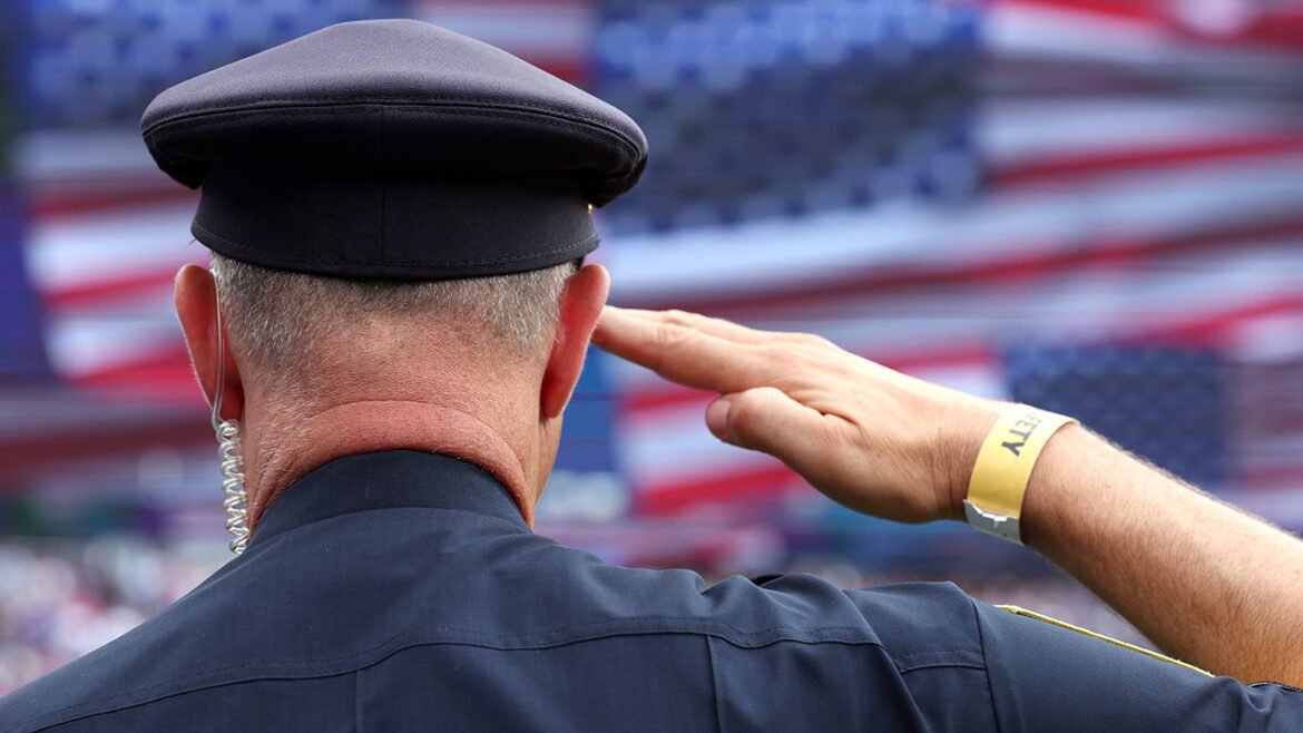 A police officer with a security wristband salutes in front of a large, blurred American flag backdrop, proudly honoring America.