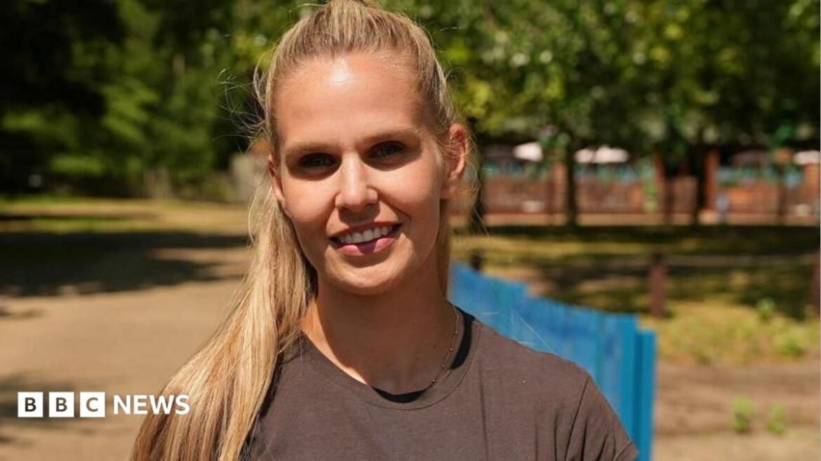 Amelia Lord is a white woman in her late 20s. She has shoulder length brown hair partly pulled back in a ponytail with frontpieces either side of her face. She has defined eyebrows and is wearing makeup, has a central nose ring and earrings, and is smiling at the camera. She wears a sleeveless black top. She is holding a pair of books and stands in front of a bookshelf with collections of books on it, including titles by Rebecca Yarros and the Harry Potter series by JK Rowling. 