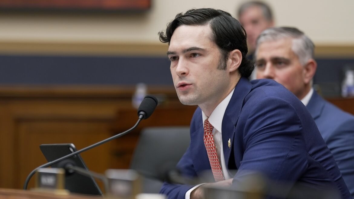 A man in a navy suit speaks into a microphone at a desk in a formal meeting room, with another man seated behind him and blurred background figures.