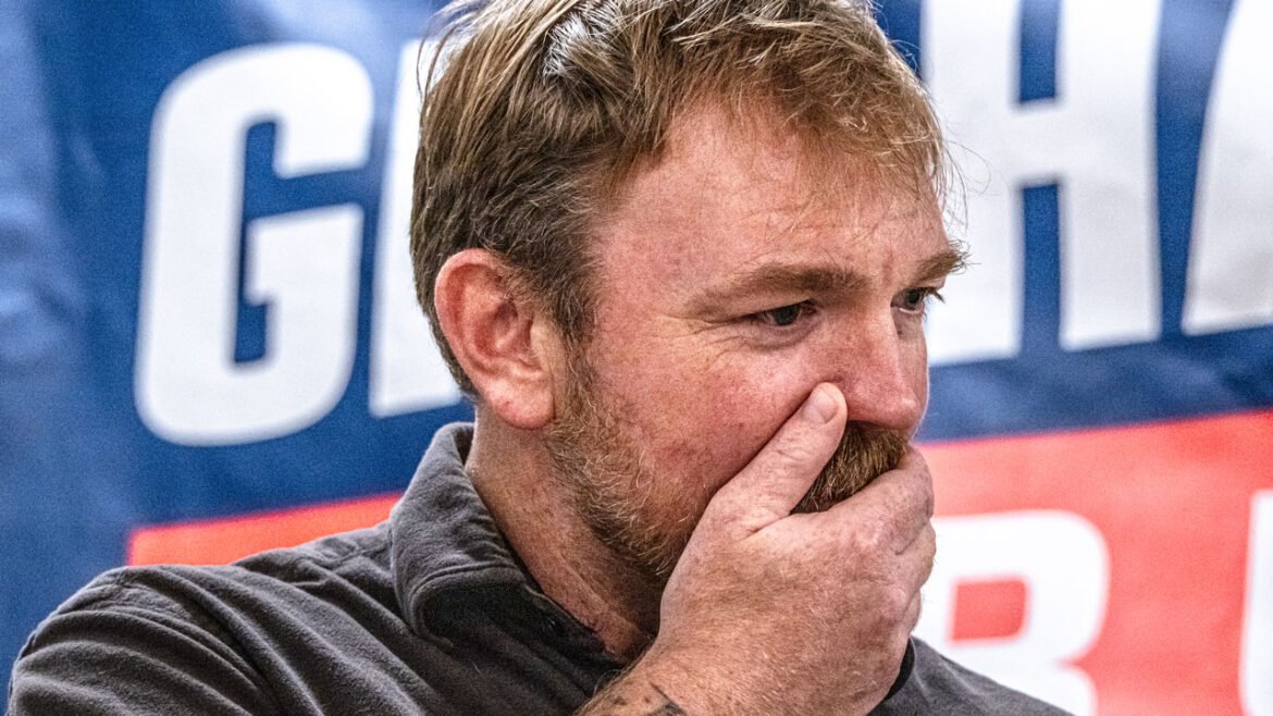 A man with light brown hair and a beard covers his mouth with his hand, appearing thoughtful or concerned. A blue banner with large white letters is blurred in the background.