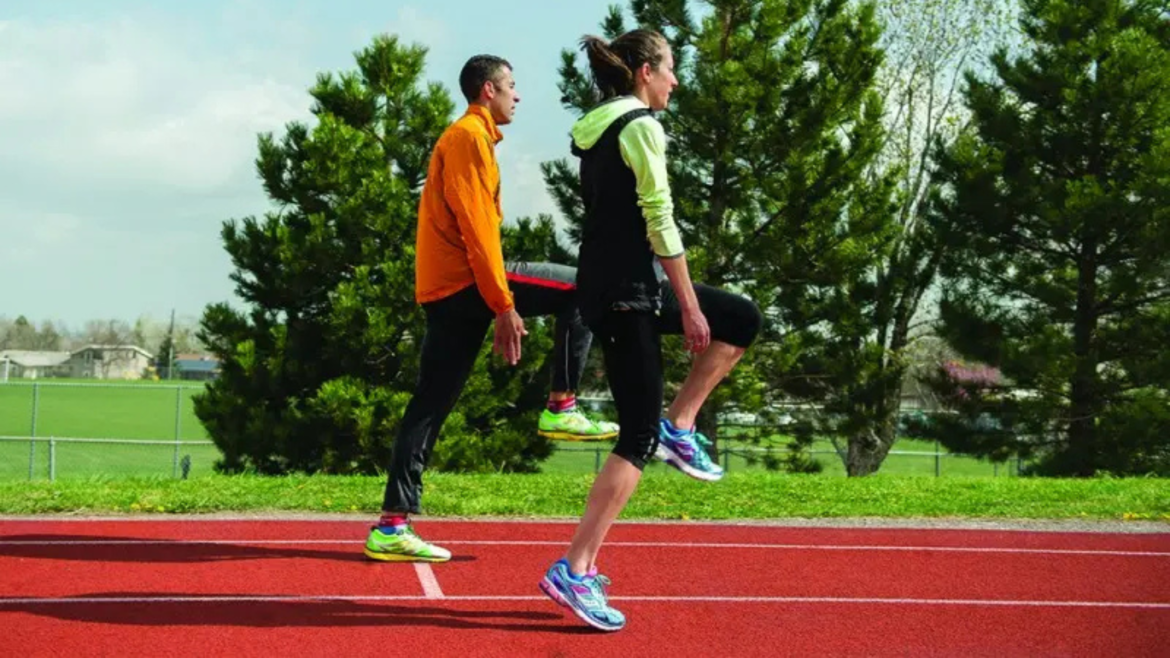 two people in running clothes on track performing under-leg clap during a-skips running drill