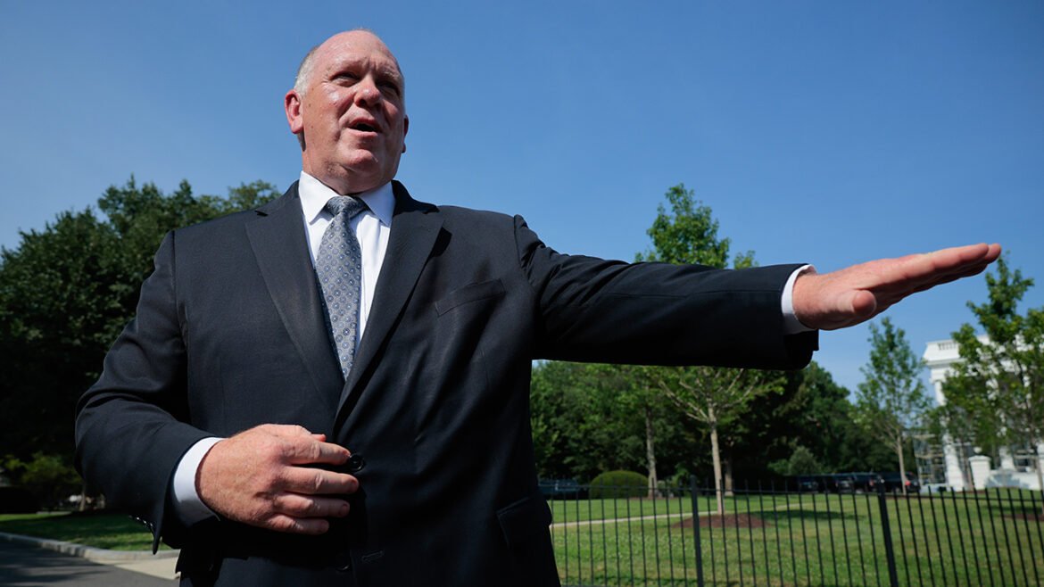 A man in a dark suit stands outdoors, gesturing with his right arm extended to the side, with green trees and a blue sky in the background.