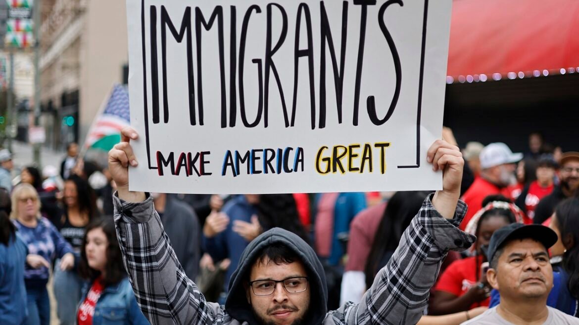 A person in a crowd holds a sign reading Immigrants make America great. People are gathered in the background, some holding flags, at an outdoor event or protest.