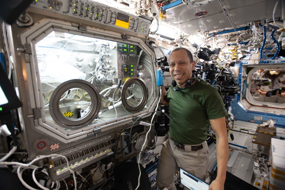 NASA astronaut Chris Williams shows off the Destiny laboratory module’s Microgravity Science Glovebox aboard the International Space Station. WIlliams was supporting semiconductor crystal research to help advance the commercial space economy and promoting Earth-based industries. He is wearing a dark green t-shirt and khaki pants with a brown belt. He is shown smiling with a headset around his neck.