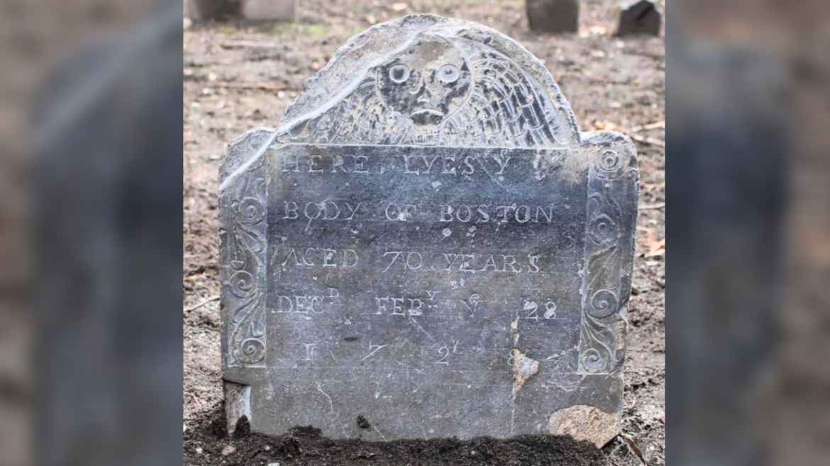 A gray gravestone in a barren landscape with a death's head decoration and inscription