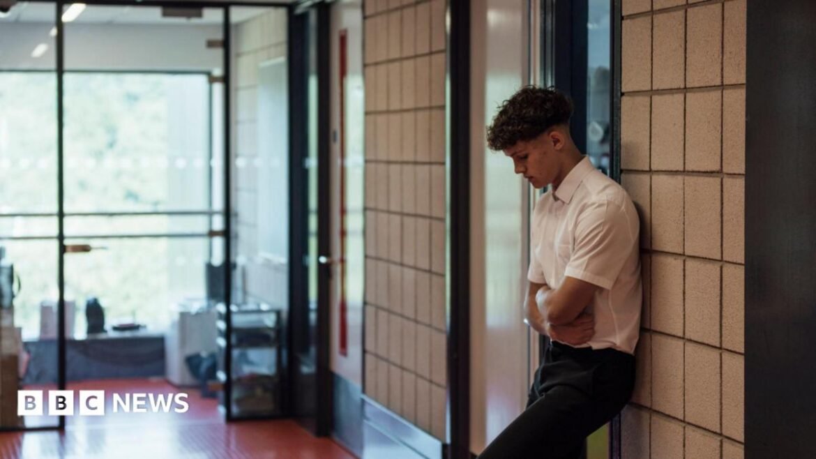 A school boy looks forlorn stood with arms folded outside a school classroom