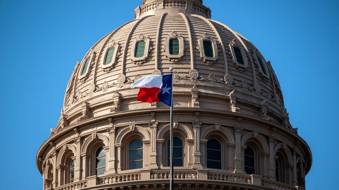 The Texas state flag waves in front of the ornate dome of the Texas State Capitol building under a clear blue sky.